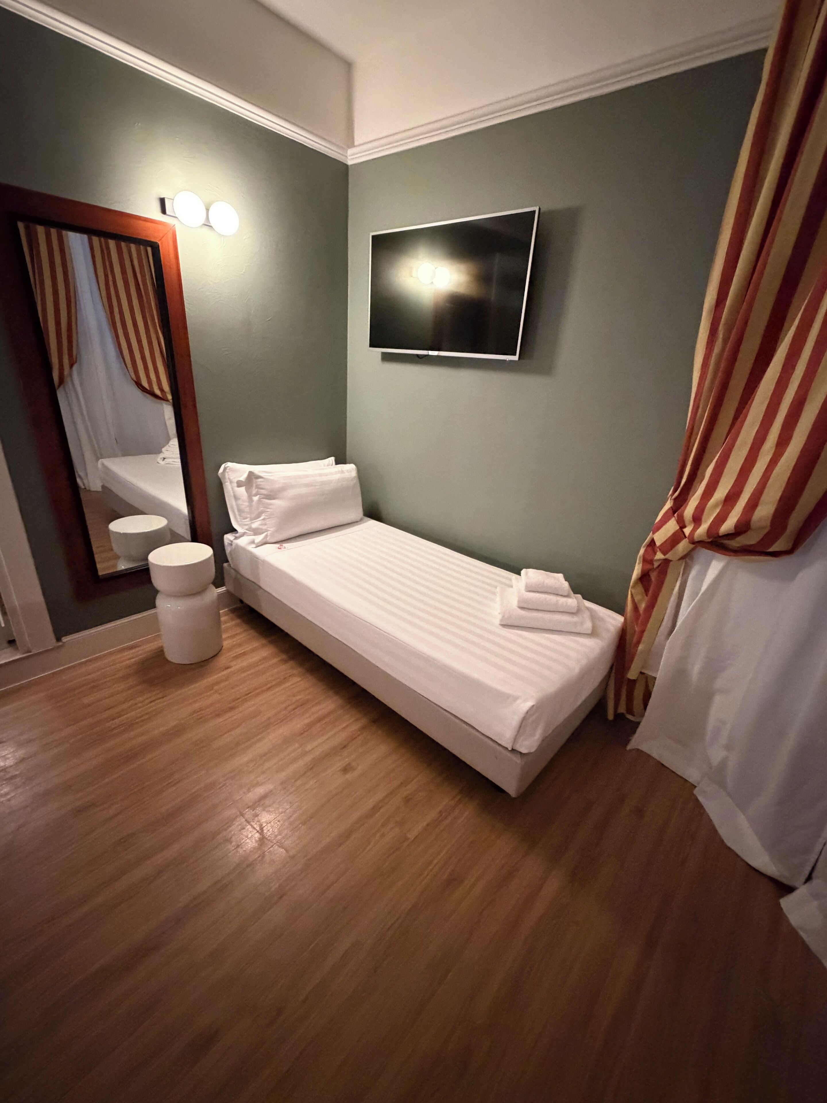 A single bed with a white mattress and pillows, a small bedside table, a wall-mounted TV, and a mirror with lights.