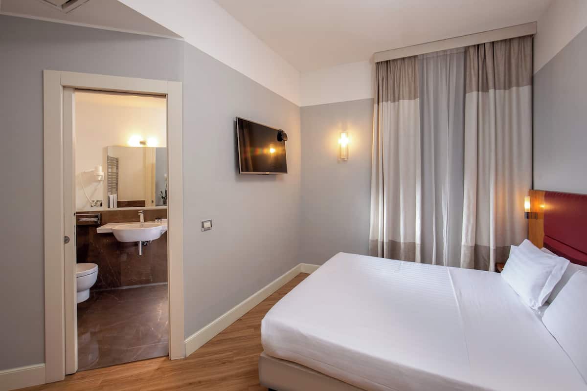 A hotel room with a bed, a bathroom with a sink and toilet, and a TV mounted on the wall.