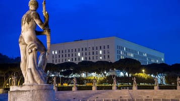 Una statua raffigura una figura con uno scettro in una piazza pubblica, con un edificio sullo sfondo.