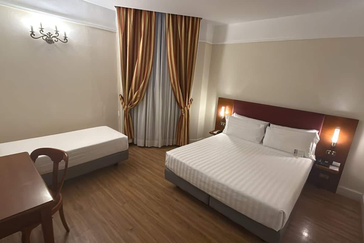 A hotel room with a bed, a desk, a chair, a nightstand, and a window with curtains.