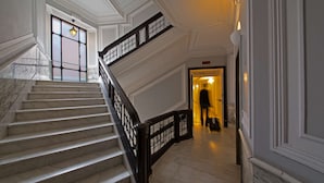 Uma escadaria imponente com degraus de mármore e corrimão preto, que leva a um corredor bem iluminado com uma porta no final.