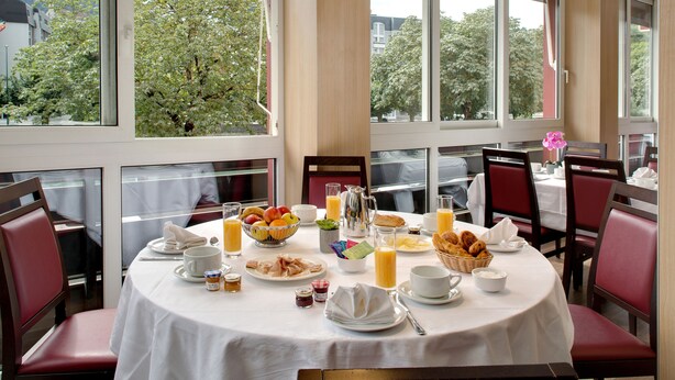 A breakfast table set with various dishes and beverages, surrounded by red upholstered chairs, in a room with large windows offering a view of trees and buildings. - greet Lourdes Centre Pyrénées (Ouverture juin 2026) (Lourdes)