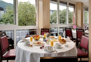 A breakfast table set with various dishes and beverages, surrounded by red upholstered chairs, in a room with large windows offering a view of trees and buildings.