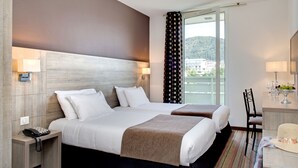 A hotel room with two beds, a desk with a lamp, a telephone, and a view of a building through a window. - greet Lourdes Centre Pyrénées (Ouverture juin 2026) (Lourdes)