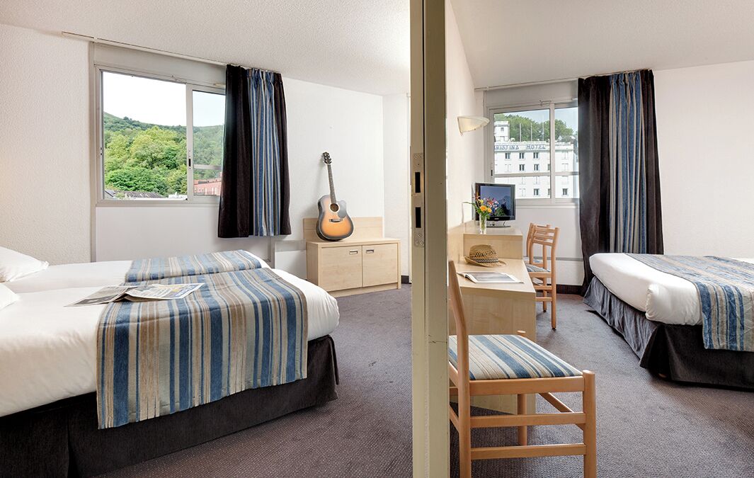 Een hotelkamer met twee bedden, een bureau, een stoel, een gitaar en uitzicht op een gebouw door het raam.