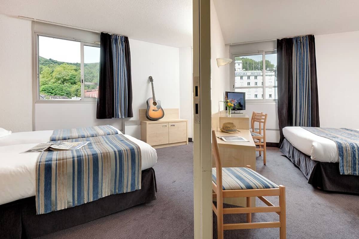 A hotel room with two beds, a desk, a chair, a guitar, and a view of a building through the window.