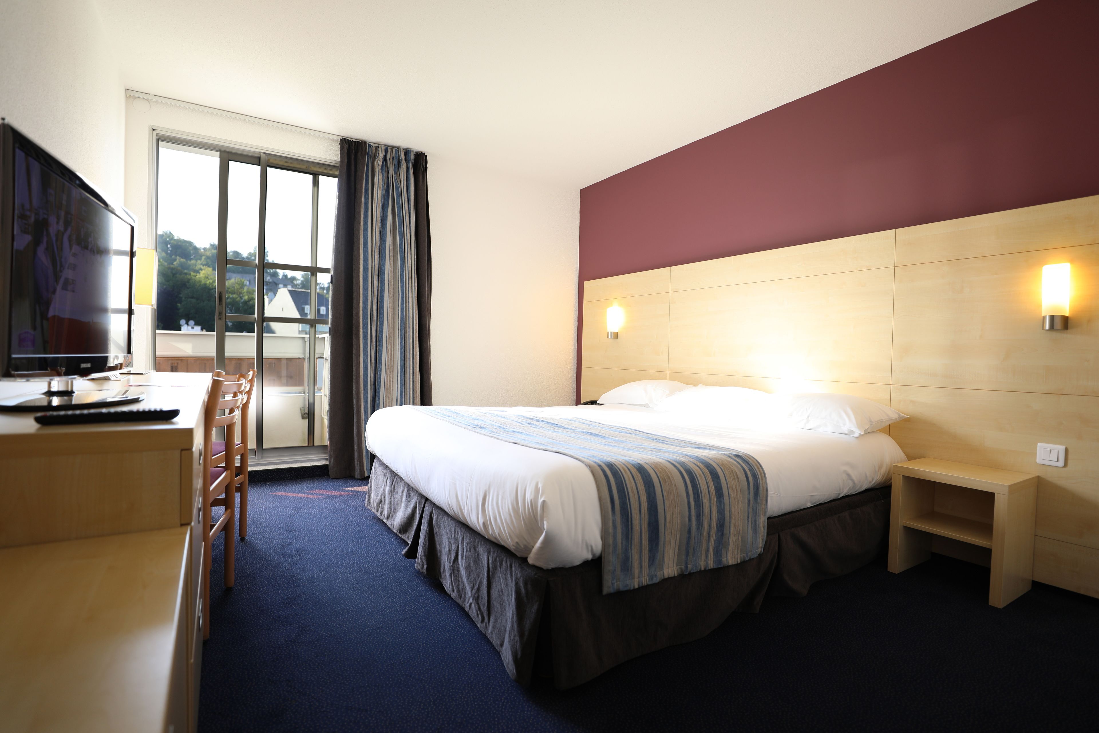 Una habitación de hotel con cama, un escritorio con silla, televisión, ventana con cortinas y mesita de noche.
