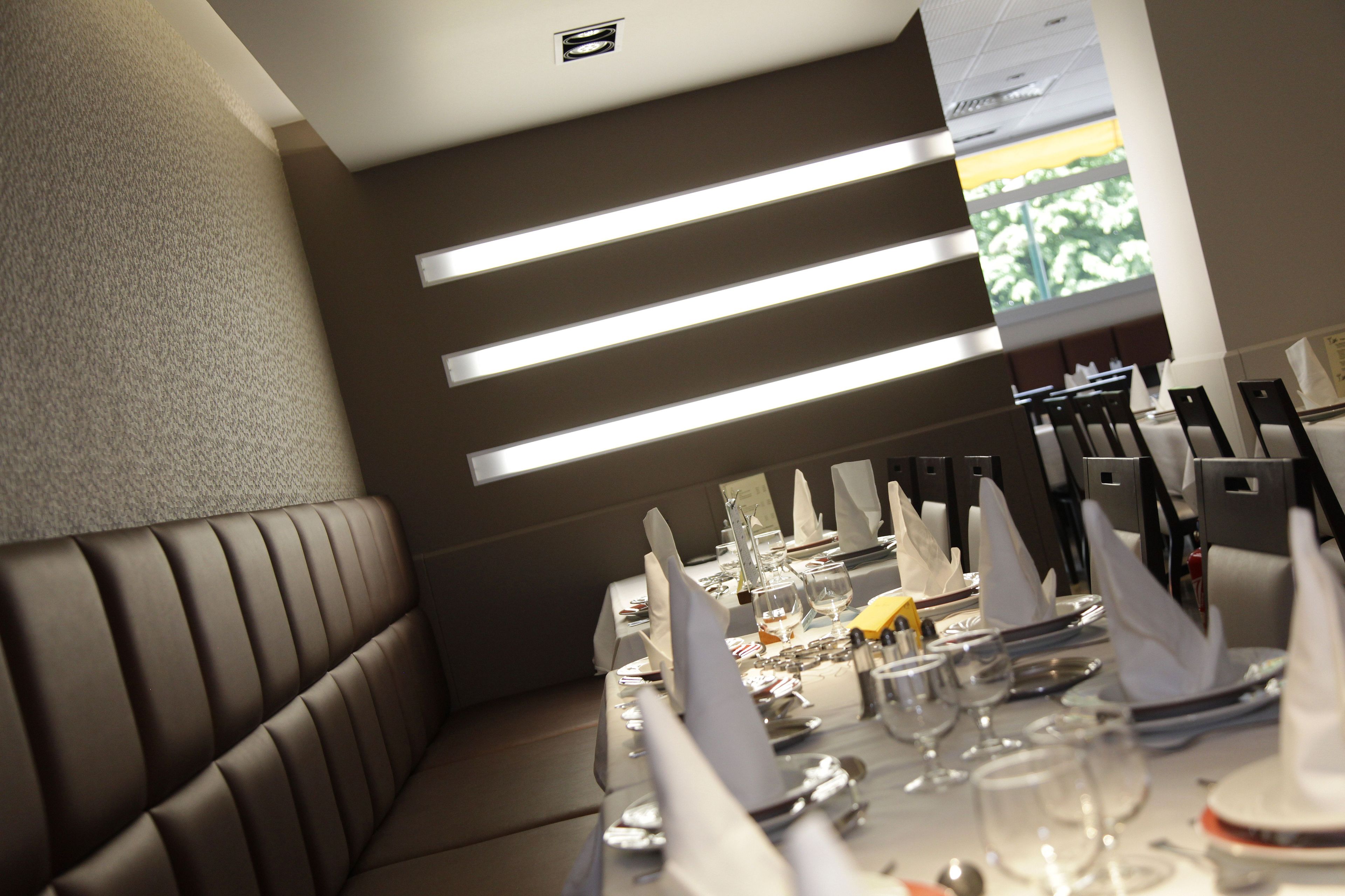 Un restaurante con una larga mesa de comedor, elegantemente dispuesta con servilletas blancas, cubiertos y copas. Sillas alineadas a lo largo de la mesa, y el ambiente presenta un diseño moderno con una pared texturizada y un elemento de iluminación en el techo.