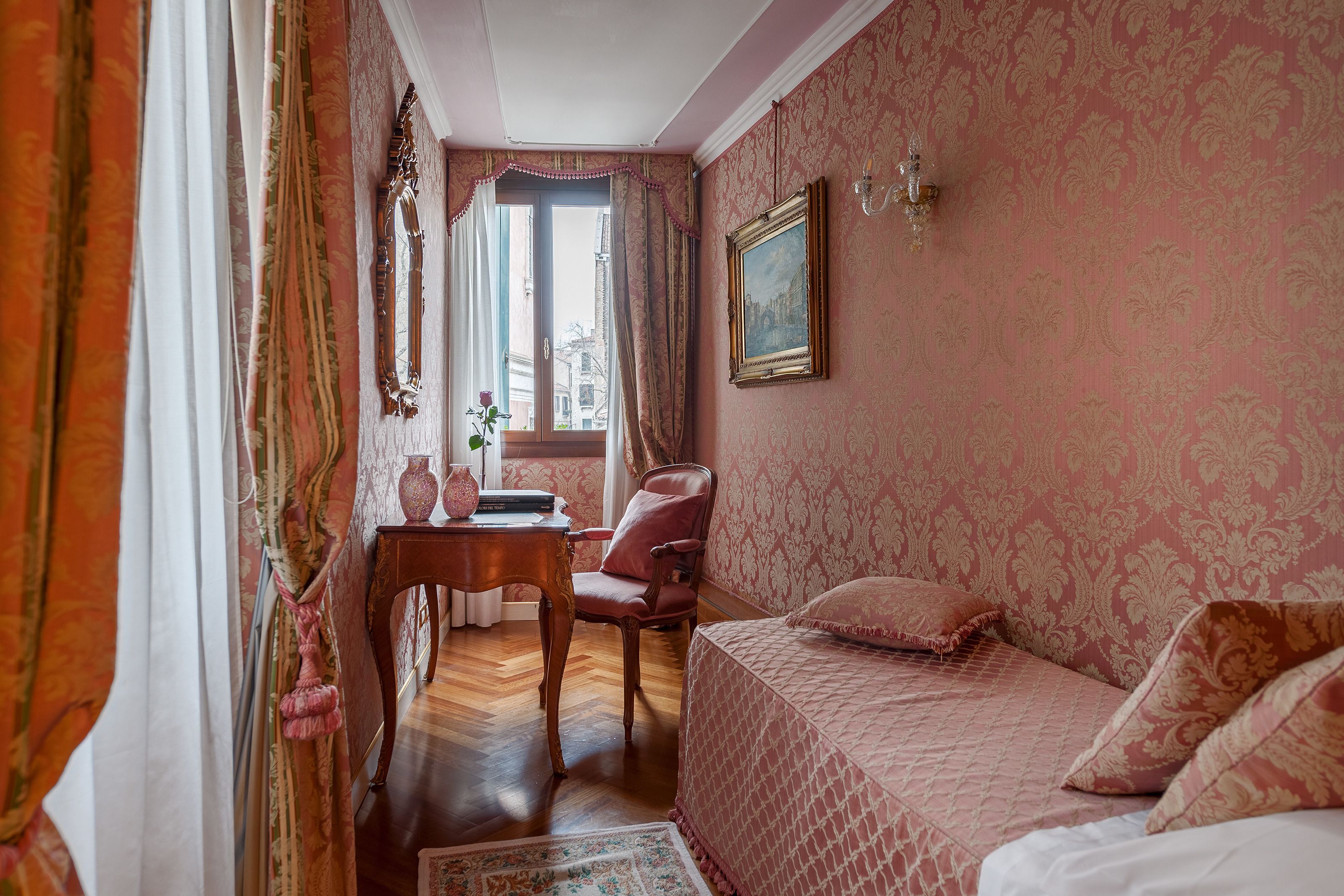 A room with a bed, a chair, a table with a vase, and a window with curtains.