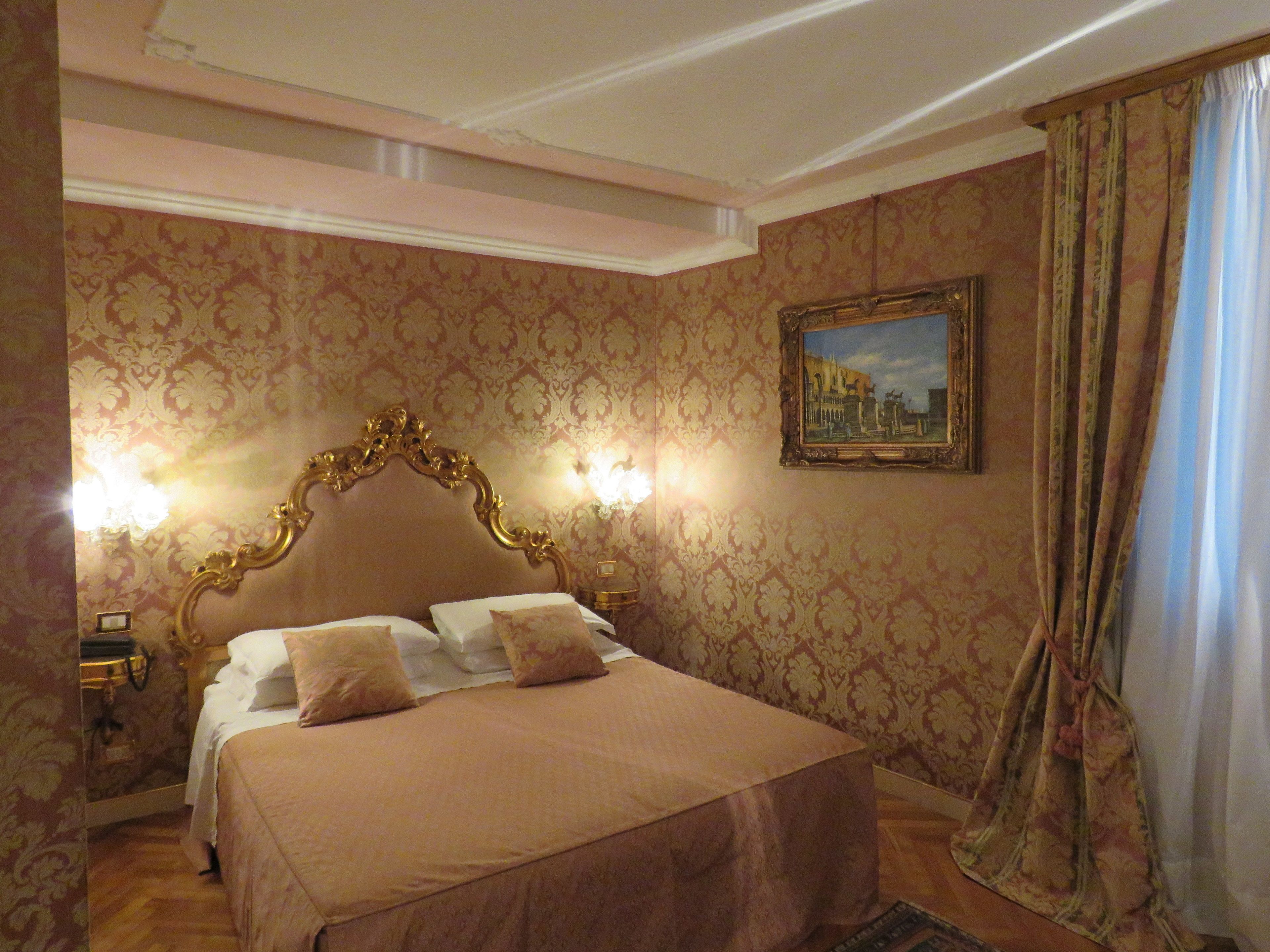 A hotel room with a large bed, ornate headboard, patterned wallpaper, and a framed painting on the wall.