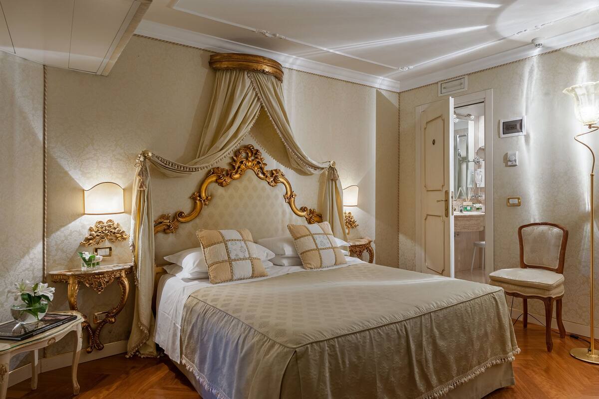 A classic four-poster bed with a canopy, a bedside table with a lamp, and a chair in a well-lit room with a wooden floor.