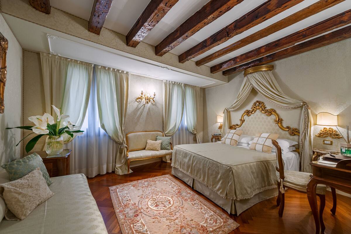 A bedroom with a four-poster bed, a sitting area with a sofa and armchair, and a large window with curtains.