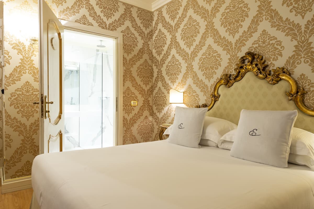 A hotel room with a large bed, ornate headboard, and patterned wallpaper.