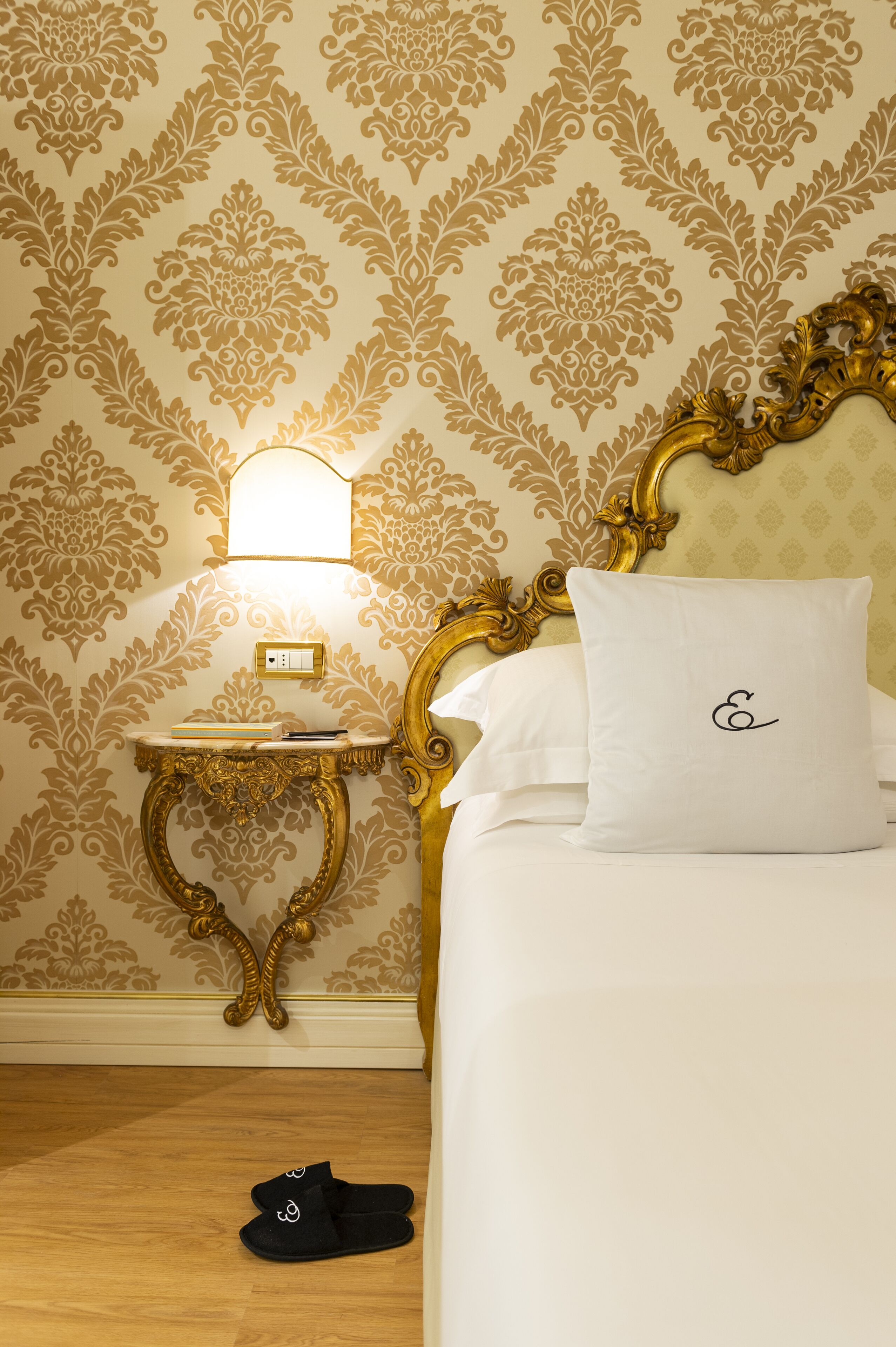 A neatly made bed with a white pillow and a gold-framed headboard. A small bedside table with a lamp and a book is visible. A pair of black slippers is placed on the floor beside the bed.