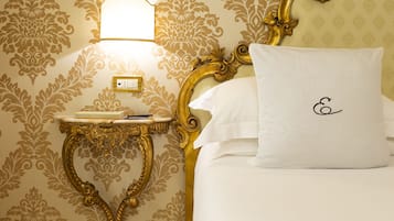 Una cama bien hecha con un almohadón blanco y un cabecero con marco dorado. Se ve una mesita de noche con una lámpara y un libro. Un par de zapatillas negras está colocado en el suelo junto a la cama.
