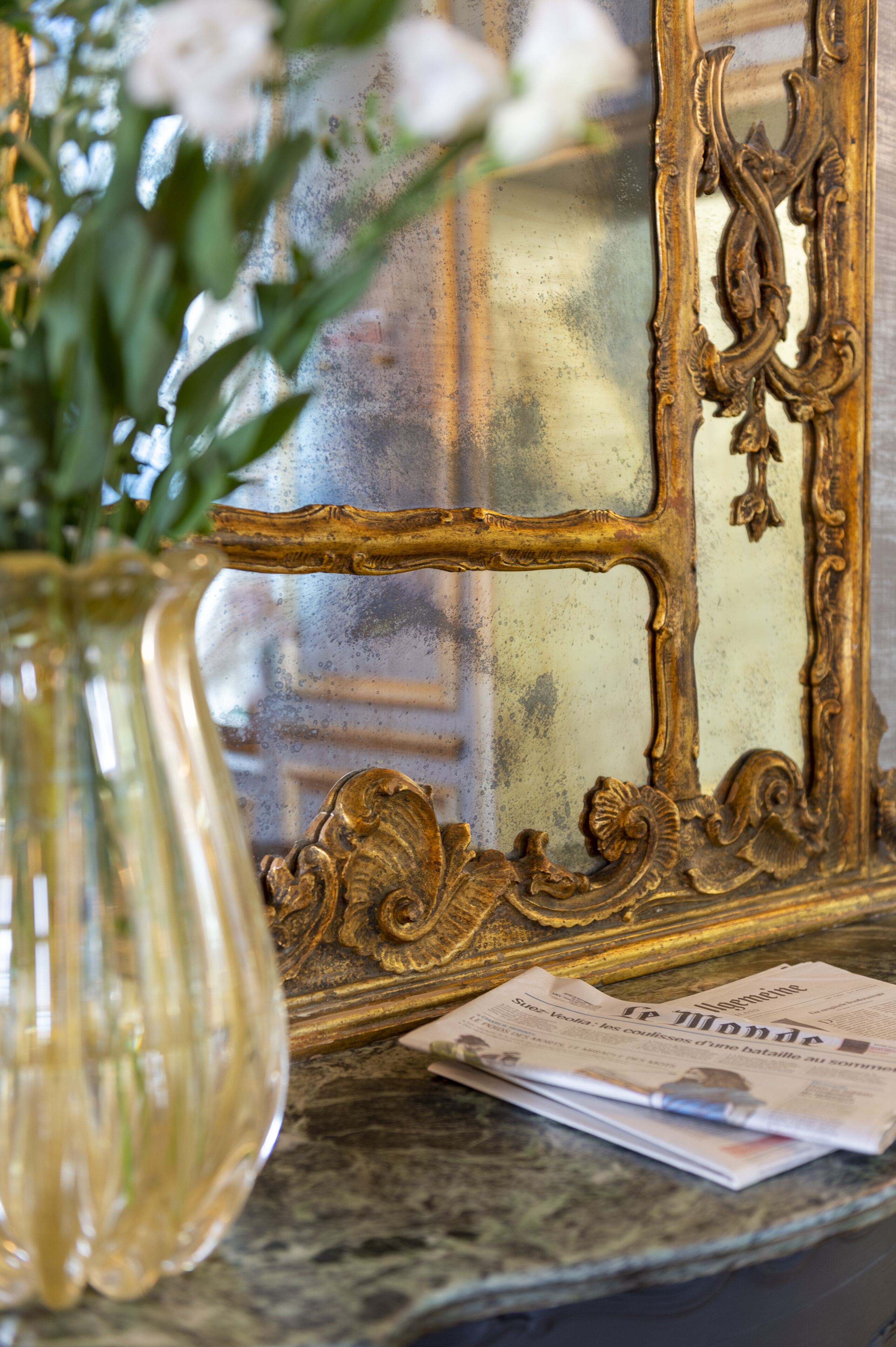A glass vase with flowers on a marble surface, a newspaper, and a gold-framed mirror.