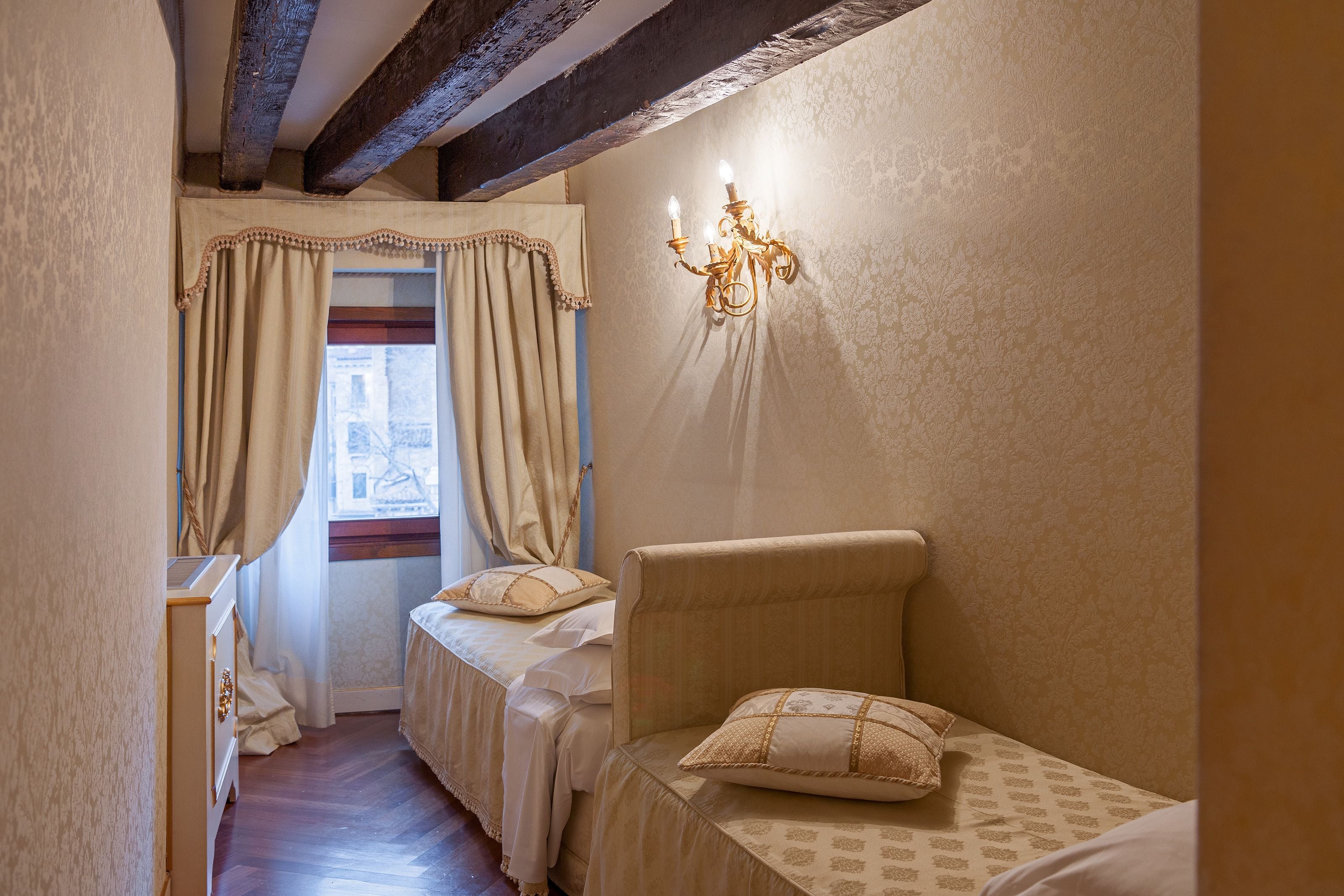 A bedroom with a bed, a nightstand, a dresser, a window with curtains, and a wall-mounted light fixture.
