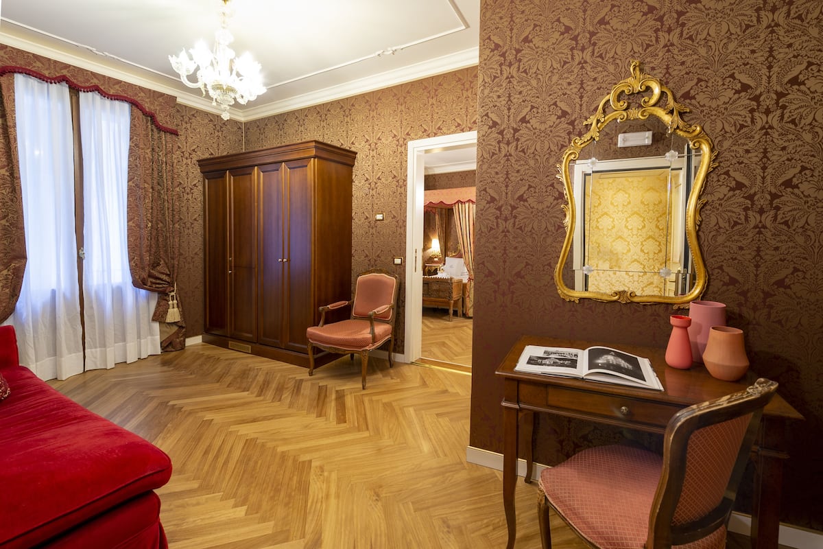 A room with a wooden floor, a red sofa, a wooden wardrobe, a chair, a desk with a mirror, and a chandelier.