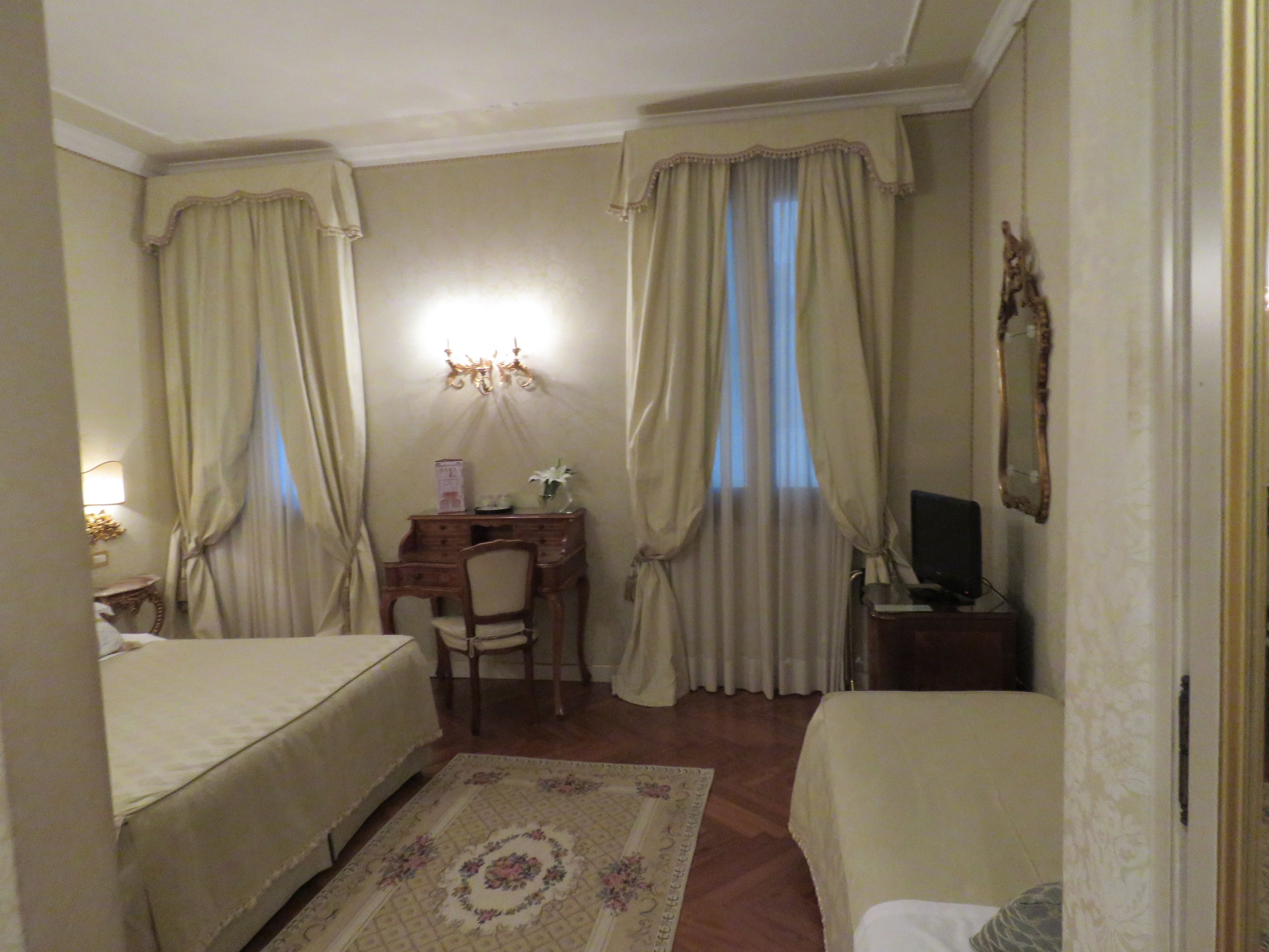 Habitación de hotel con cama, escritorio con silla, televisor y espejo en la pared.