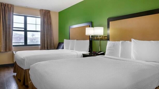 A hotel room with two beds, a large window with curtains, a bedside lamp, and a view of the cityscape. - Extended Stay America Select Suites - Detroit - Roseville (Roseville)