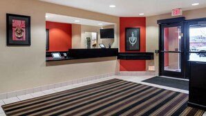 A hotel reception area with a reception desk, a large mirror, and a red accent wall. There are two exit signs above the door. - Extended Stay America Select Suites - Detroit - Roseville (Roseville)