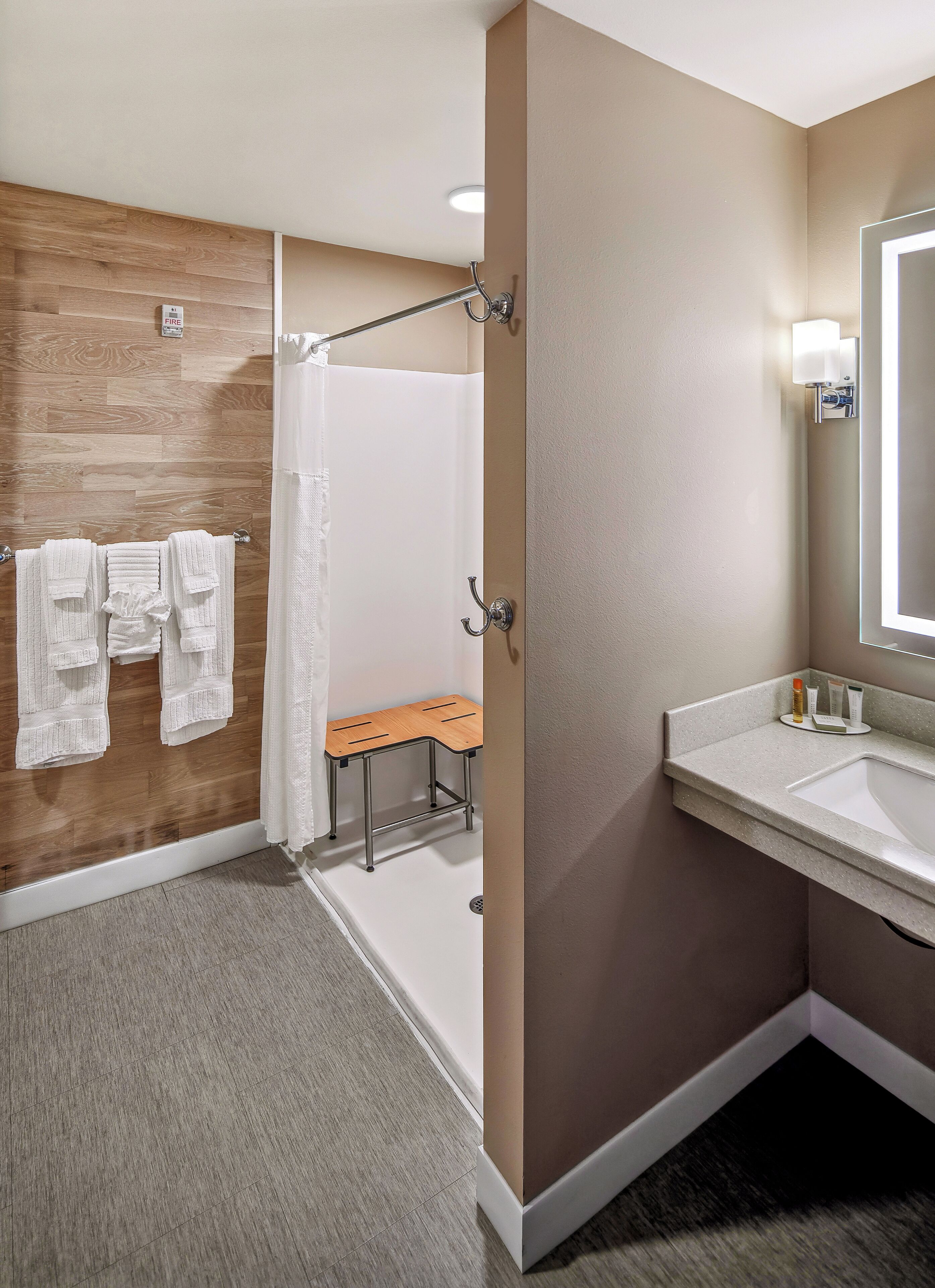 Ein modernes Badezimmer mit einer Wandpaneele aus Holz, einer Dusche mit Glastür, einer Sitzbank und einem Waschtisch mit Spiegel und Pflegeprodukten.