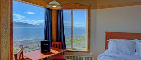 Habitación de hotel con un ventanal que ofrece vistas al mar y a las montañas, un escritorio de madera con una silla, cama con ropa blanca y un ventilador de techo.