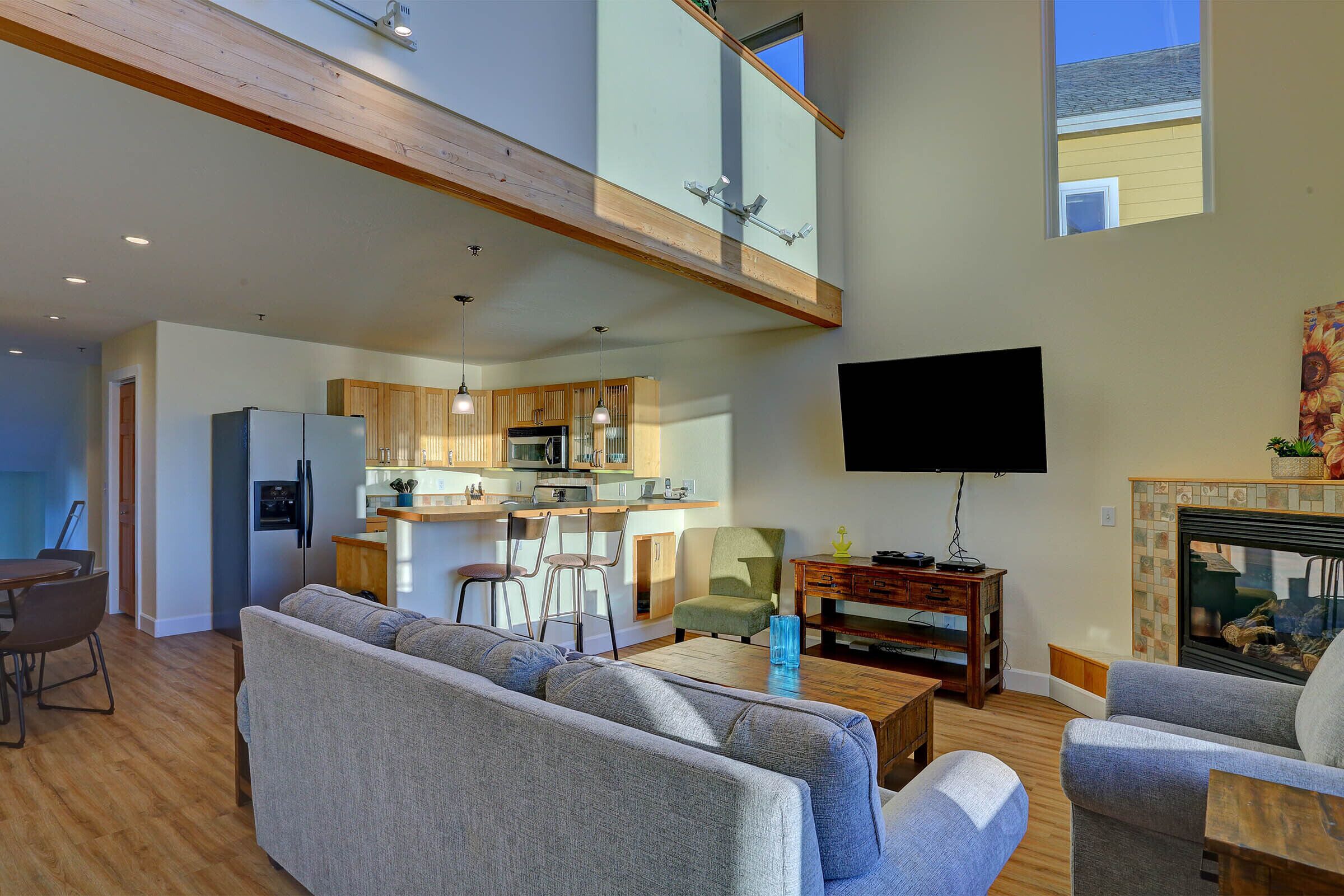 A spacious living area with a high ceiling, a kitchen with wooden cabinets, a flat-screen TV mounted on the wall, and a fireplace.