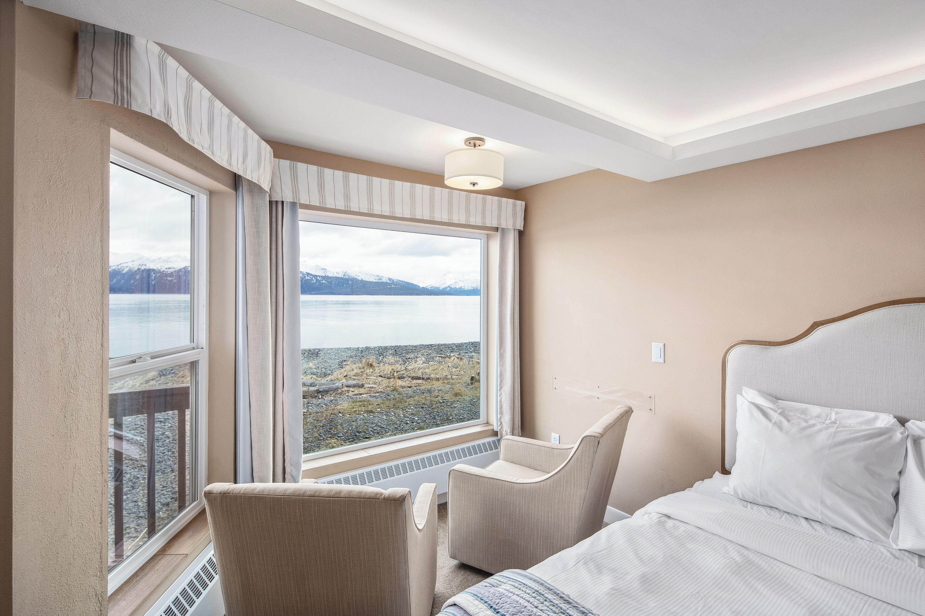 Habitación de hotel con una cama, una silla y un amplio ventanal con vistas a un lago y montañas.