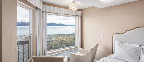 A hotel room with a bed, a chair, and a large window offering a view of a lake and mountains.