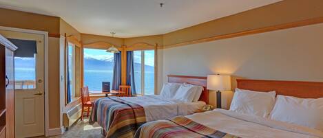 A hotel room with two beds, a desk, and a chair. The room has a view of the ocean and mountains.
