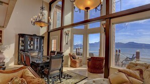 Ein geräumiges Wohnzimmer mit großem Fenster mit Blick auf ein Gewässer und Berge. Es gibt ein Flügel, bequeme Sitzgelegenheiten und eine Kronleuchter.