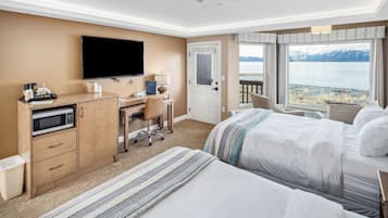 A hotel room with two beds, a desk with a computer, a TV, and a view of a lake with mountains.