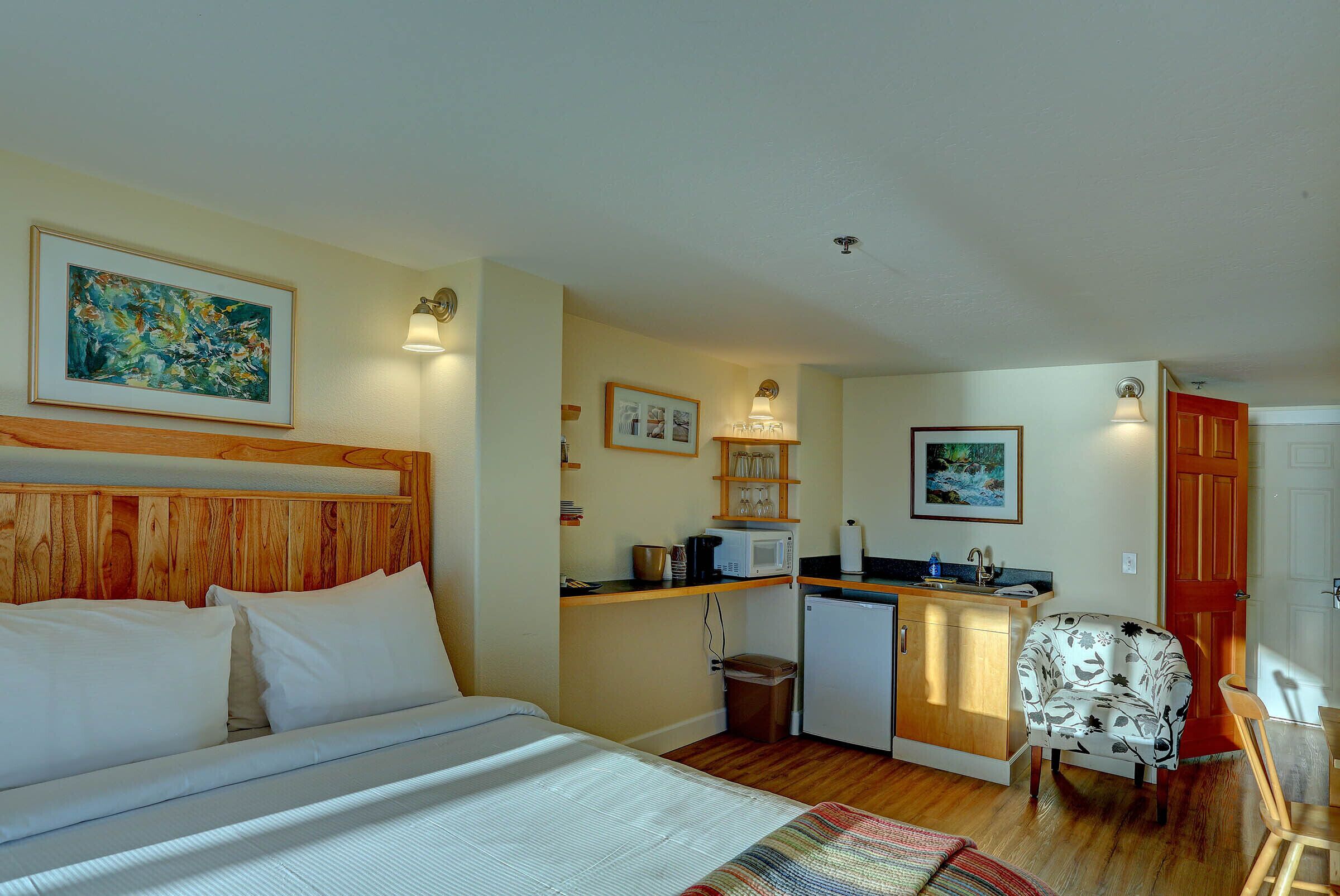 A bedroom with a wooden headboard, a bed with white linens, a small kitchenette with a microwave, and a chair.