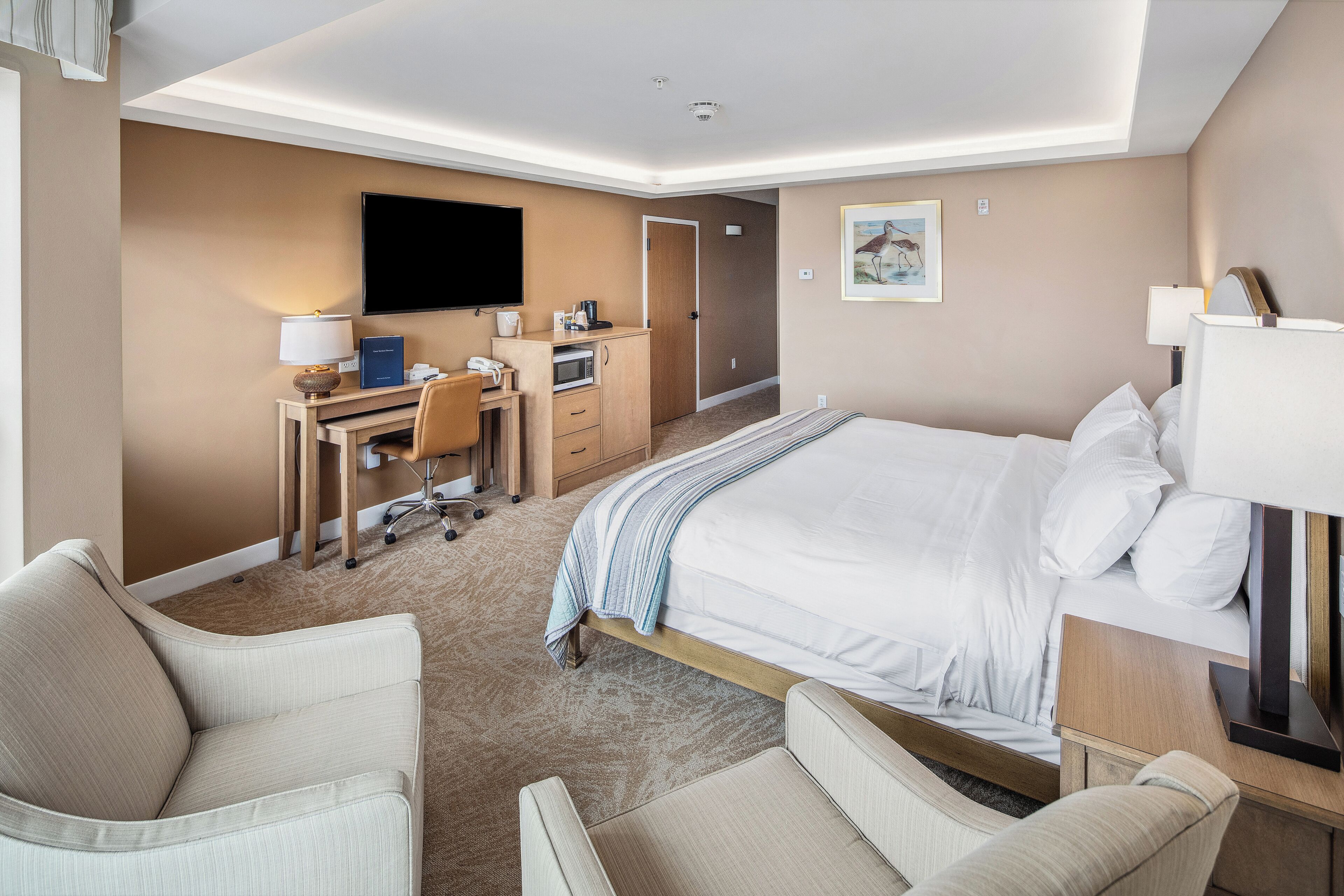 Ein Hotelzimmer mit einem Bett, zwei Sesseln, einem Schreibtisch mit Computer, einem Fernseher und einem Bild an der Wand.