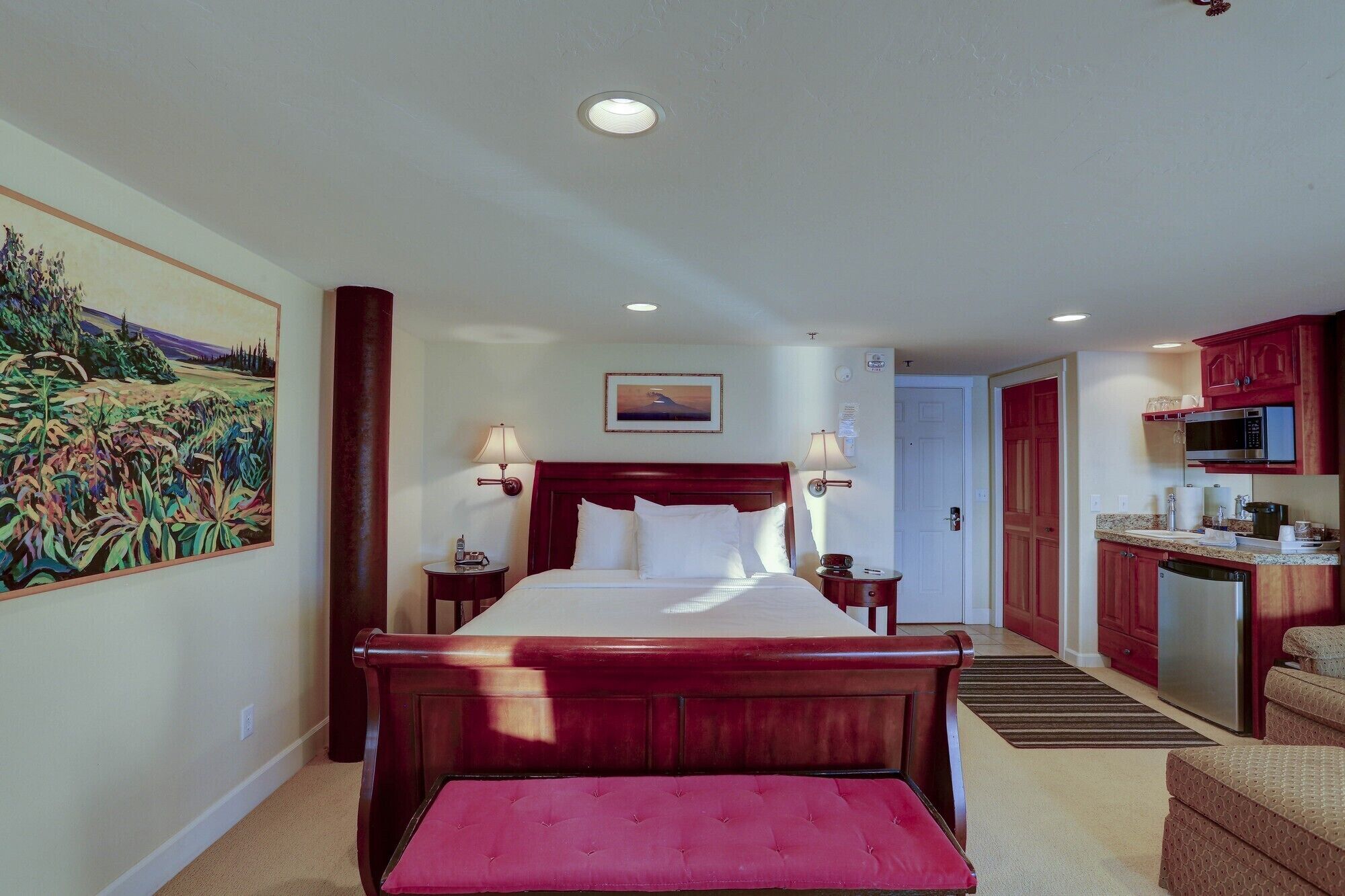 Une chambre d’hôtel avec un grand lit, une petite table de chevet, une commode, un tableau au mur, une porte donnant sur la cuisine et un banc dans un coin.