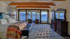 Ein Schlafzimmer mit einem Bett, einer Bank, einem Schminktisch und einem großen Fenster mit Blick auf Berge und Wasser.