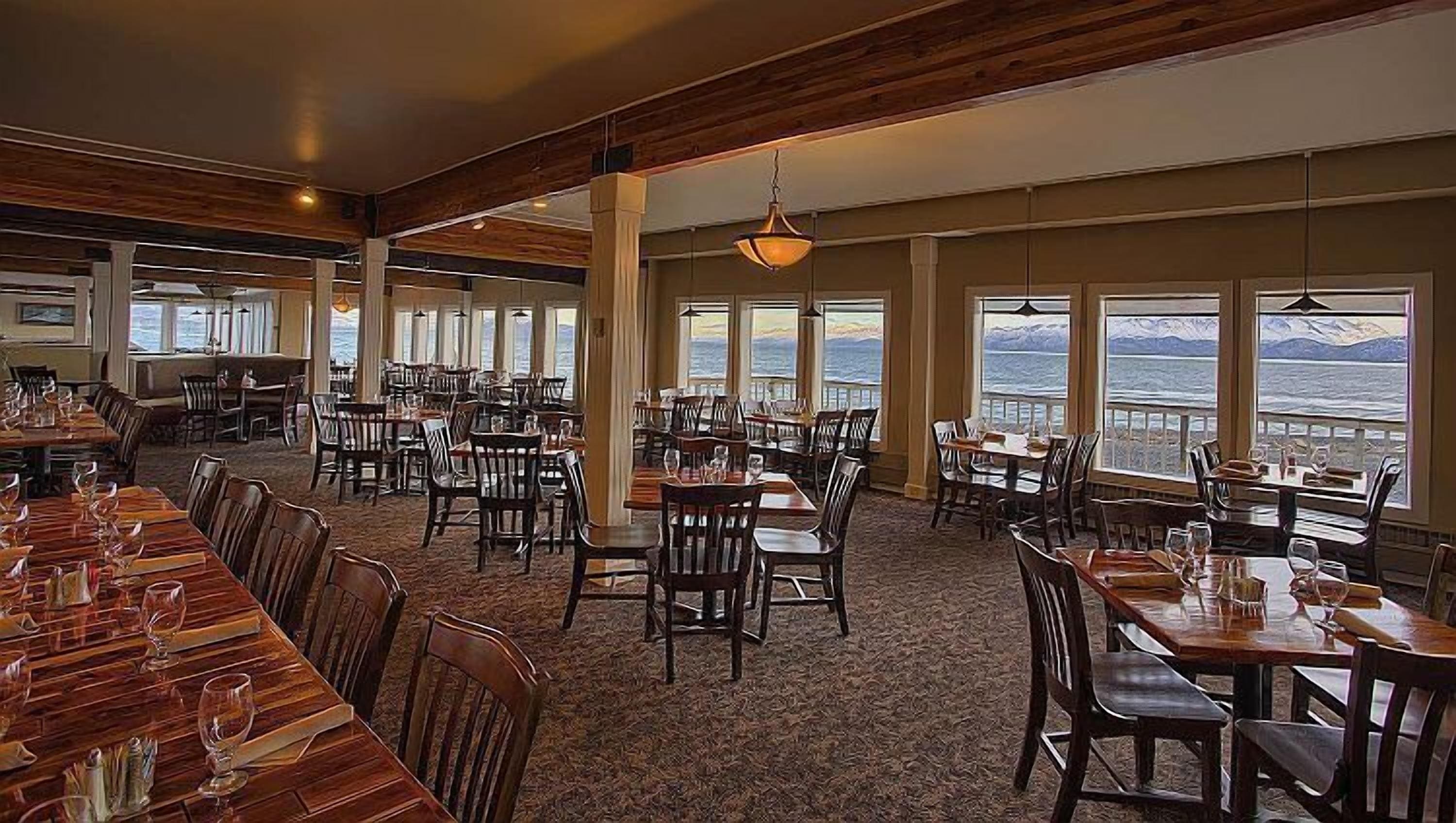 Un'ampia sala da pranzo con numerosi tavoli e sedie, ampie finestre vista mare e montagne, e travi a vista in legno al soffitto.