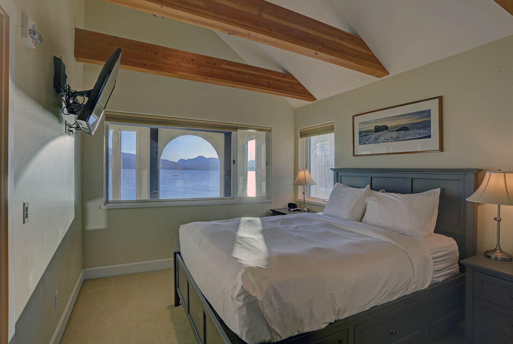 A bedroom with a bed, a nightstand, a lamp, a picture on the wall, and a window with a view of the sea.