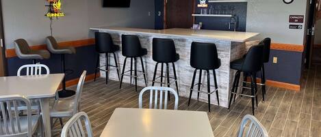 A bar area with a counter, stools, and a neon sign. There are tables and chairs around the bar.