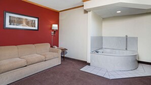 Habitación de hotel con un sofá color beige, un cuadro en la pared, una mesita de noche con una lámpara y una bañera en esquina.