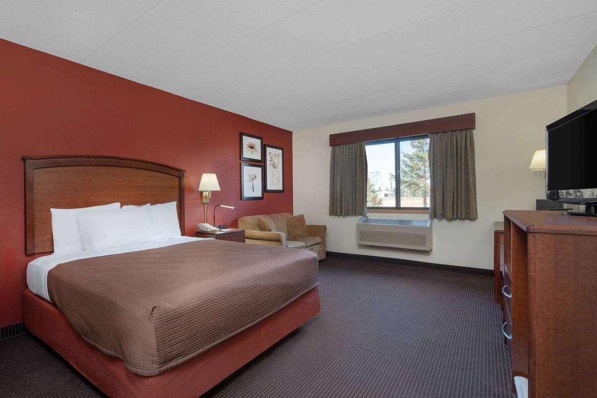 A hotel room with a large bed, a desk with a lamp, a chair, an air conditioning unit, a television, and a window with curtains.