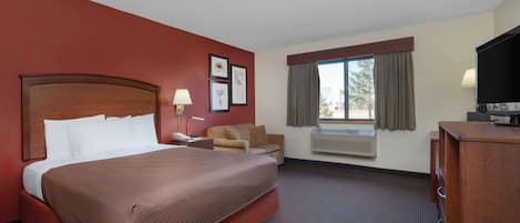A hotel room with a large bed, a desk with a lamp, a chair, an air conditioning unit, a television, and a window with curtains.