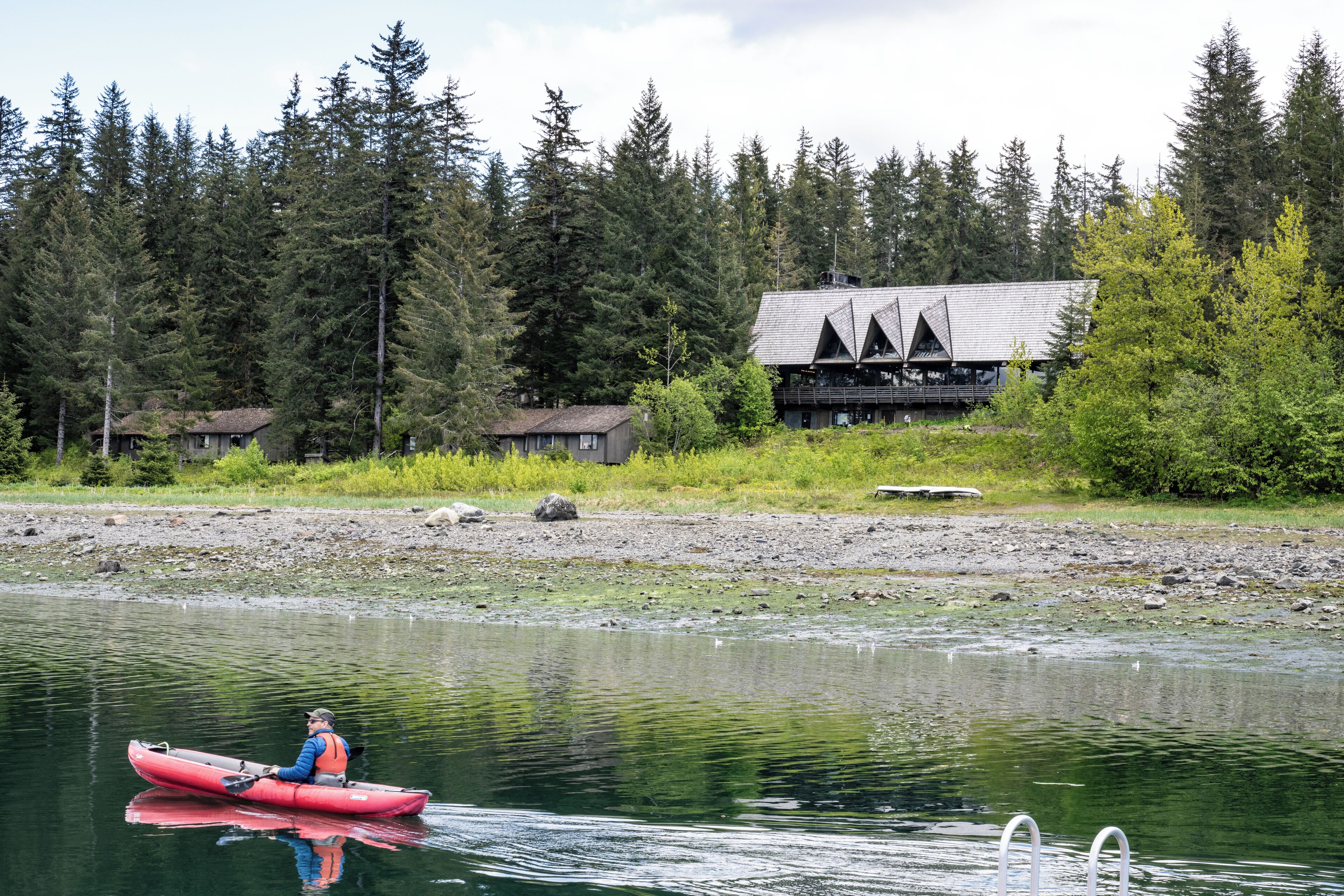 A person in a red kayak on a calm river, surrounded by dense forest and a lodge with a unique triangular roof.