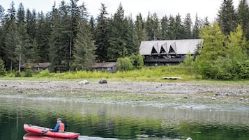 A person in a red kayak on a calm river, surrounded by dense forest and a lodge with a unique triangular roof.