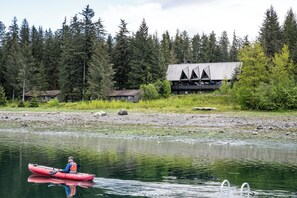 A person in a red kayak on a calm river, surrounded by dense forest and a lodge with a unique triangular roof. - Glacier Bay Lodge (Gustavus)