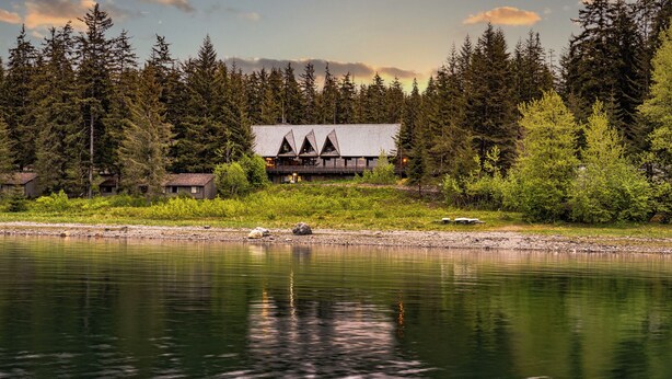 A lodge with a modern design, situated by a lake with dense forest in the background. - Glacier Bay Lodge (Gustavus)