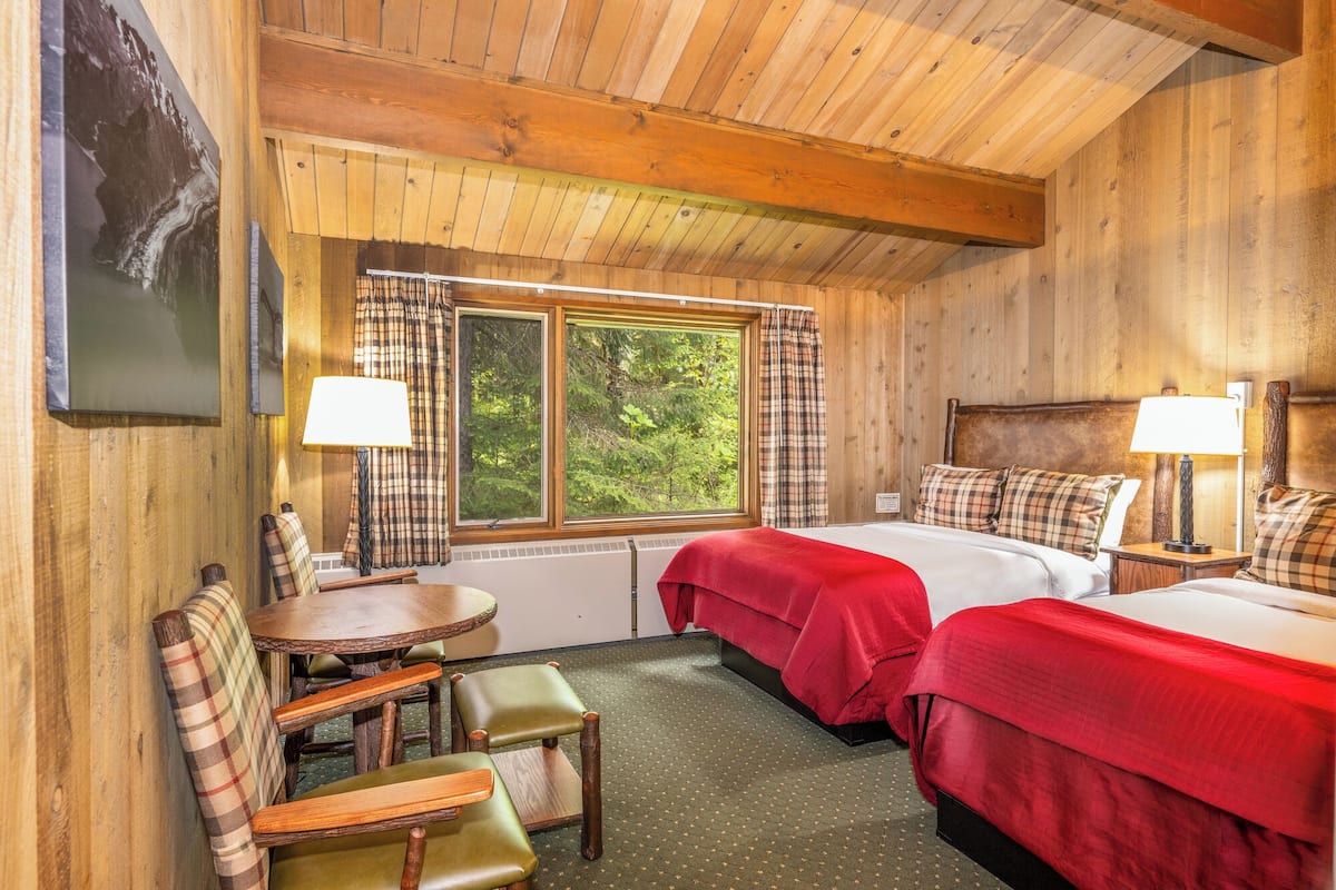 A wooden cabin room with two beds, a table, chairs, and a window with curtains.