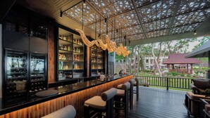 Una zona de bar con taburetes de madera, una barra con estantes llenos de botellas y vistas al área exterior con una terraza de madera y un edificio de techo rojo al fondo.
