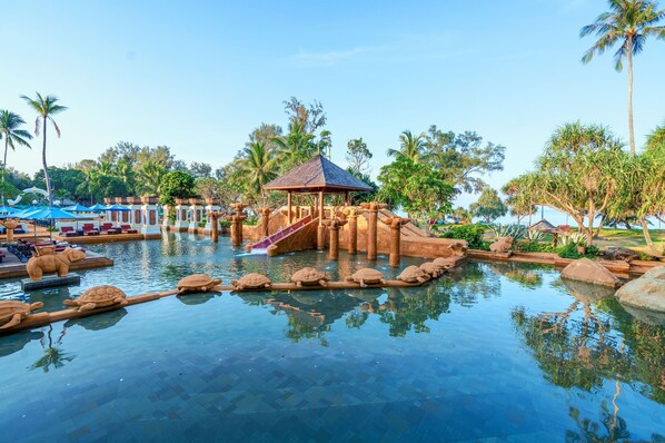 Zona de piscina con puente de madera, palmeras y un pabellón.